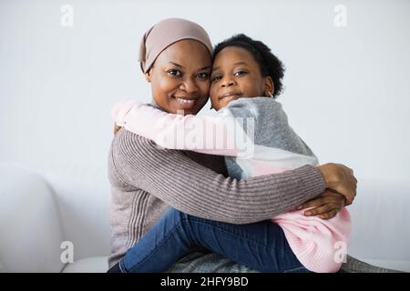 Bambina femmina che abbraccia la sua piacevole madre musulmana mentre si siede insieme su un comodo divano su sfondo bianco studio. Famiglia felice sorridendo e guardando la macchina fotografica. Foto Stock