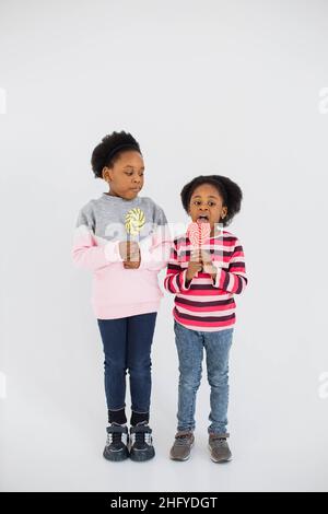 Carino afroamericana sorelle vestito casual leccare dolce colorful lollipops mentre in piedi su sfondo bianco studio. I bambini felici godono di caramelle di zucchero al coperto. Foto Stock