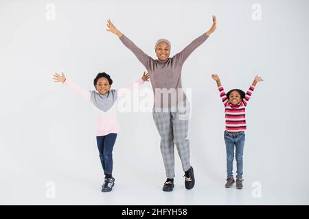 Bella donna musulmana afroamericana con due simpatiche figlie che alzano le mani e sorridono sulla macchina fotografica mentre si trova in studio. Concetto di famiglia, felicità ed emozioni. Foto Stock