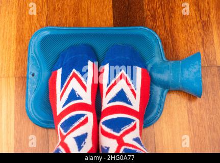 Persona che indossa i calzini Union Jack mantenendo i piedi caldi sulla bottiglia di acqua calda. Aumento di energia, elettricità, prezzi del gas, costo della vita... concetto. Foto Stock