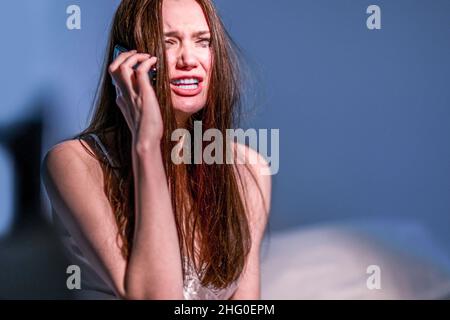 Una donna con un volto sporco da mascara si siede a letto e piange, parlando al telefono. Depressione. Ottieni le cattive notizie. Cuore rotto. Per soffrire. Smettiti. Foto Stock