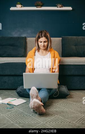 Donna caucasica in auricolari guardare lo schermo del laptop guardare seduto sul pavimento mentre si prende corso lontano o sul portatile a casa. Vista frontale. Ragazza Foto Stock