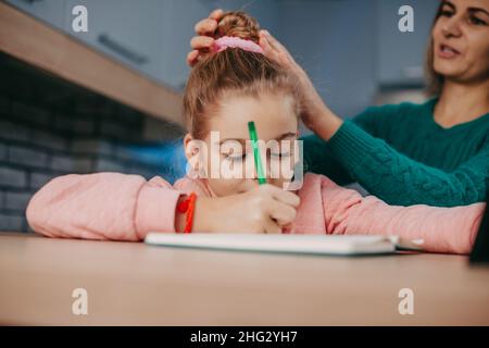 Ragazza caucasica che scrive nel suo taccuino mentre sua madre fa il suo aspetto buono. Madre che aiuta il lavoro della figlia, disegno grande per tutti gli scopi. Bambini Foto Stock