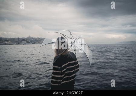 Primo piano della vista posteriore della giovane donna caucasica con capelli biondi e maglione nero che tiene un ombrello trasparente e guarda il mare in una giornata piovosa Foto Stock