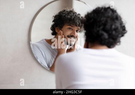 Attraente uomo indiano bearded che tocca faccia che applica moisturizer sul viso, in piedi vicino allo specchio nella stanza da bagno moderna Foto Stock