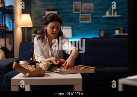 L'impiegato d'ufficio a casa ha cenato prendendo una fetta di pizza calda dalla scatola di cartone sul tavolo con deliziosi hamburger e patatine fritte. Donna che ha un menu di fast food da asporto e birra di fronte alla televisione. Foto Stock