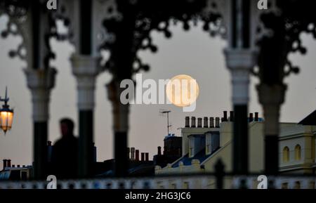 Brighton UK 18th gennaio 2022 - il Wolf Moon affonda dietro il lungomare di Brighton e il banco di bandstand presto questa mattina . Tradizionalmente la prima luna piena dell'anno è conosciuta come la Luna del lupo . Credit Simon Dack / Alamy Live News Foto Stock