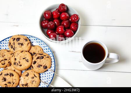 Tazza bianca con caffè, biscotti tradizionali sul piatto e ciliegie dolci in una ciotola di ceramica su sfondo bianco di legno, primo piano. Colazione gustosa, morni Foto Stock