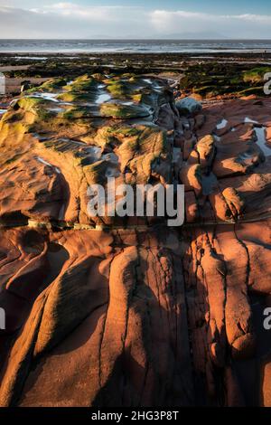 Luce serale su rocce di arenaria esposte a bassa marea alla spiaggia di Maryport sulla costa Cumbria, Inghilterra, con il Solway Firth alle spalle Foto Stock