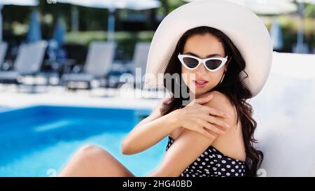 donna brunetta con cappello di paglia e occhiali da sole che applicano protezione solare sul braccio vicino alla piscina Foto Stock