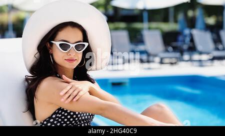 giovane donna con cappello di paglia e occhiali da sole che applicano protezione solare sul braccio vicino alla piscina Foto Stock