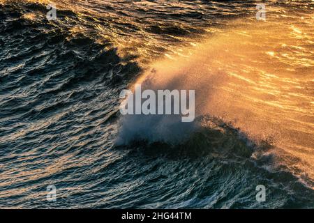 Spray soffiato dalla cresta di un'onda da forte vento e retroilluminato da un tramonto dorato. Foto Stock