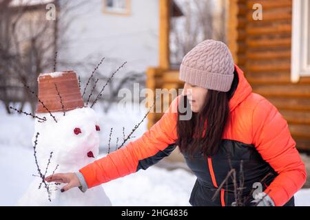 Donna di età media da lei stessa facendo pupazzo di neve sul cortile di casa di giorno mentre la neve sta cadendo giù con la casa di legno sullo sfondo. Genitori Foto Stock
