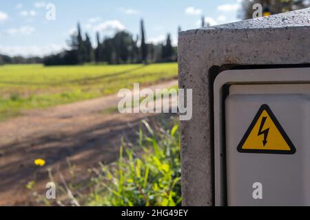 Primo piano di un segnale di pericolo elettrico. Sullo sfondo, fuori fuoco, una campagna mediterranea in una giornata di sole Foto Stock