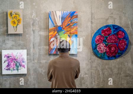 Vista posteriore dell'ospite africano maschile della galleria d'arte in piedi di fronte al muro con esposizioni e guardando uno di loro Foto Stock