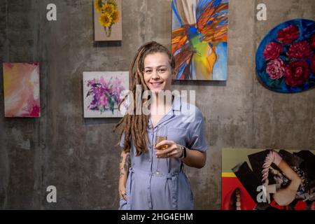 Felice giovane donna con dreadlock tostare con flute di champagne mentre si trova di fronte alla macchina fotografica in galleria d'arte Foto Stock