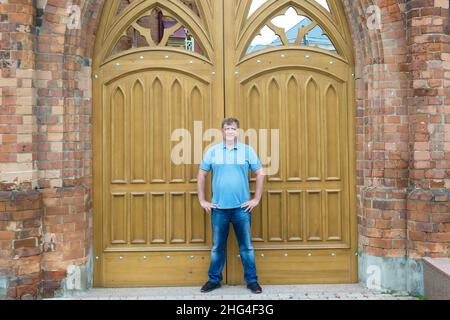 l'alto uomo biondo di 40-45 anni sorge all'alto cancello di legno di un edificio in mattoni. Foto Stock