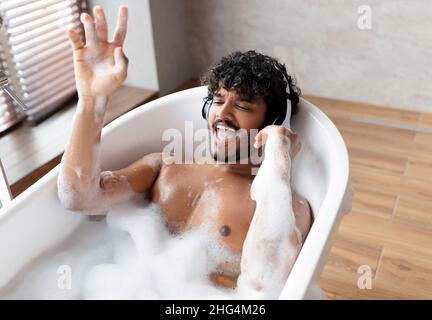 Ragazzo indiano gioioso in cuffie wireless godersi la musica e cantare mentre si trova in bagno caldo bolle a casa Foto Stock
