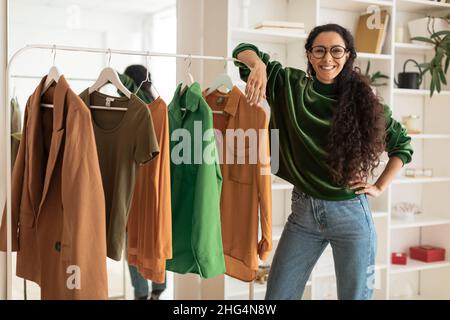 Happy Female Designer in piedi vicino Clothing Rail in Modern Showroom Foto Stock