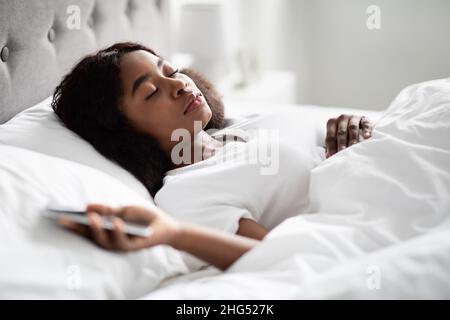 Donna nera infelice che stesa a letto, tenendo il cellulare Foto Stock