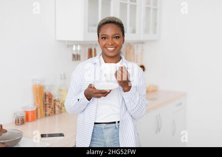 Allegro bella millennial donna nera in abiti casual bevendo la sua bevanda calda preferita in tazza bianca Foto Stock