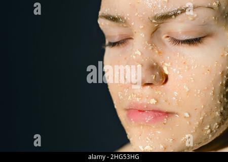 Faccia della ragazza con maschera cosmetica di farina d'avena con miele. Prevenzione dell'acne negli adolescenti. Volto della donna con gli occhi chiusi su sfondo scuro. Foto Stock