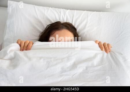 Vista dall'alto di una giovane donna che nasconde il viso sotto la coperta mentre si trova in un letto confortevole. Foto Stock