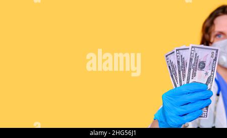 Un medico mantiene uno stipendio in dollari. Un'infermiera con soldi per il lavoro e una maschera protettiva su sfondo giallo, spazio per la copia del banner Foto Stock