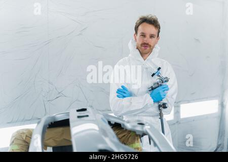 Ritratto di un giovane pittore di auto con una pistola in una camera di vernice spray. Lavoratore di sesso maschile in garage auto riparazione Foto Stock