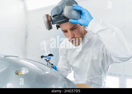 Il pittore di auto professionista nella cabina di verniciatura toglie la sua maschera protettiva per verificare la qualità del lavoro svolto. Pittore automatico durante il lavoro Foto Stock