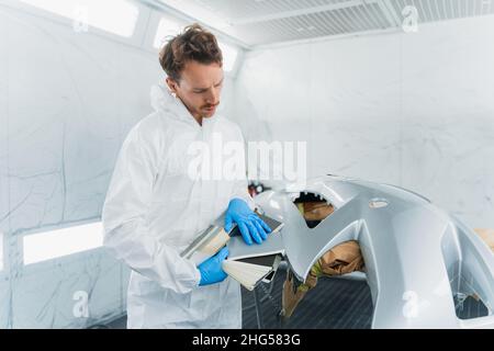 Il giovane lavoratore maschile di un negozio di riparazione carrozzeria in una cabina di verniciatura auto confronta il colore di un paraurti auto con i campioni Foto Stock