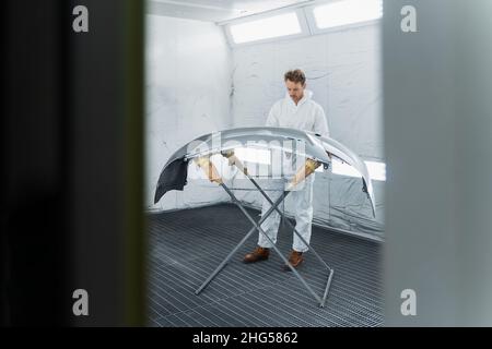 Il pittore auto maschio in un vestito bianco protettivo funziona con un paraurti in una cabina di verniciatura auto Foto Stock