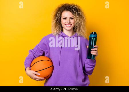 Foto di giovane bella ragazza sportiva tenere acqua bere fresco allenamento tenere palla isolato su sfondo di colore giallo Foto Stock