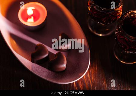 Praline al cioccolato a forma di cuore con liquore alla ciliegia in due bicchieri con rosa rossa e candele in un ambiente romantico. Concetto di San Valentino. Romantico. Foto Stock