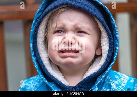 Vista ravvicinata sul volto grimace di un bimbo sconvolto che esplica in lacrime durante un'esplosione emotiva. Ragazzino infelice che indossa il cappuccio blu all'aperto. Foto Stock