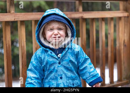 Primo piano ritratto di un ragazzo di tre anni che indossa una giacca blu con cappuccio esterno durante l'inverno avere un tantrum. Sconvolto e piangere con la faccia scrunched. Foto Stock