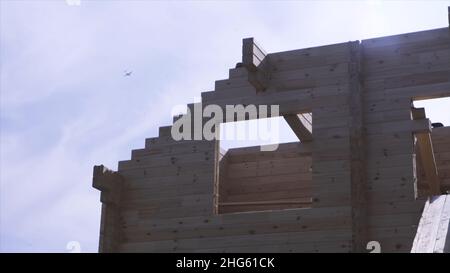 Struttura di edificio, casa di legno in legno su sfondo cielo blu con un aereo che vola via. Costruzione in corso di una casa di legno contro cielo nuvoloso Foto Stock