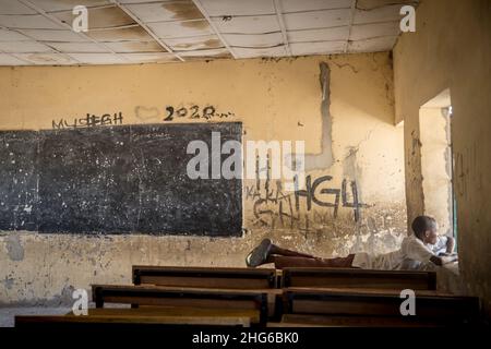 Un ragazzo ha visto adagiarsi su una scrivania in un'aula vuota della scuola elementare di Moduganari.la Nigeria nordorientale sta vivendo una rivolta dal 2009, che ha portato a 2,4 milioni di persone sfollate e circa la metà degli studenti scolastici deve lasciare l'istruzione. Foto Stock