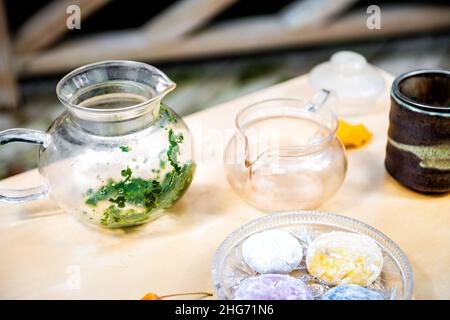 Foglie di tè verde in teiera di vetro con colorata torta di riso mochi dessert giapponese daifuku su tavola di legno ancora vita closeup Foto Stock