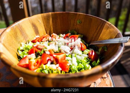 Primo piano di insalata fresca in ciotola di legno con lattuga, pomodori rossi e cipolle tagliate a fette con fondo bokeh di terrazza all'aperto in garde Foto Stock