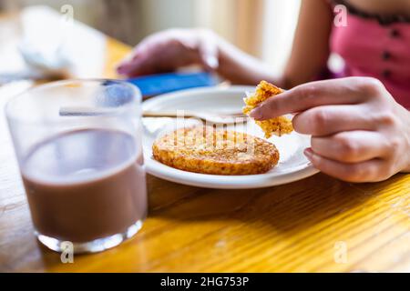 Macro closeup di donna seduta al tavolo della colazione che tiene mangiare patate fritte hash browns con mano su piatto da bicchiere di latte al cioccolato e sfondo Foto Stock