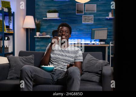 Giovane uomo seduto sul divano guardando la televisione guardare film di intrattenimento godendo di attività di lesiure a casa dopo una giornata impegnativa. Ragazzo che tiene popcorn ciotola bere birra durante la serie commedia Foto Stock