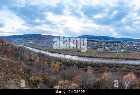 Stretto fiume che passa oltre l'insediamento tra la campagna autunnale contro alte montagne lontane sotto il cielo cupo con le nuvole piovose Foto Stock
