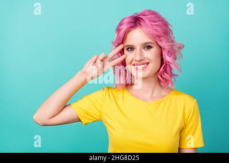 Foto di millennial dolce rosa hairdo donna mostra v-segno indossare giallo t-shirt isolato su sfondo teal Foto Stock