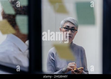 Senior Asian Company Manager brainstorming nella sala riunioni mentre ha appunti appiccicosi sul vetro in ufficio. Concetto di cooperazione e di diversità del lavoro di squadra Foto Stock