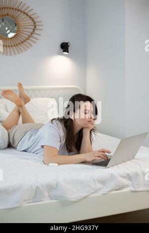 Giovane donna che usa un computer portatile mentre si trova a letto in camera da letto a casa Foto Stock