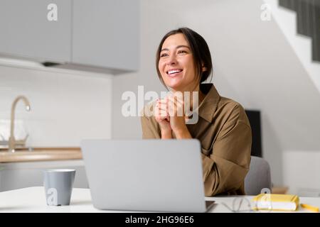 Sorridente giovane allegra donna asiatica che si gode il caffè del mattino mentre si lavora sul portatile, seduto alla scrivania in ufficio. Donna d'affari felice che fa una pausa caffè Foto Stock