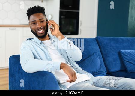 Tranquillo e bello ragazzo tranquillo che parla sullo smartphone mentre si siede sul comodo divano in elegante appartamento. Ritratto di uomo multirazziale durante la conversazione telefonica Foto Stock