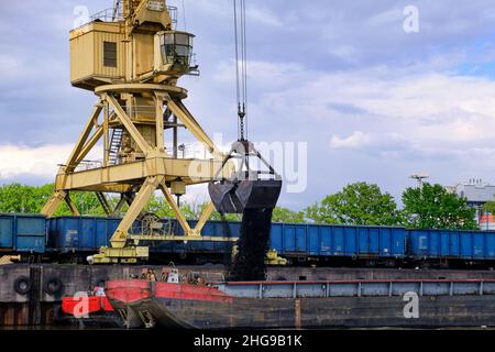 Gru fluviale portuale con benna o graniglia caricando carbone al fiume trascinare barche o chiatte ormeggiate dal molo in giorno nuvoloso. Energia, minerali, ambiente Foto Stock