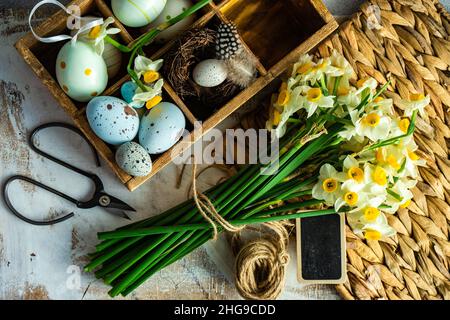 Vista dall'alto dei fiori di narciso su un tavolo con intemperie accanto ad una scatola di uova di Pasqua dipinte Foto Stock